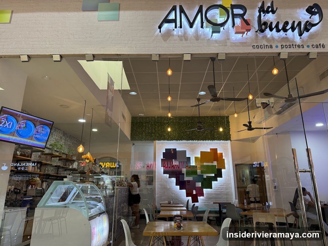 A view through the glass entrance doors of a cafe named 'AMOR al bueno'. The interior features white brick walls, wooden tables, and colorful geometric art. A large TV screen displays images of food, and a glass display case is visible on the left.