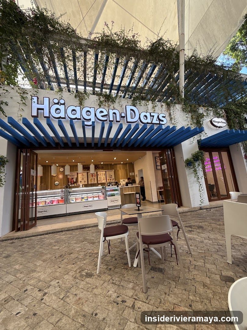 An outdoor view of a Häagen-Dazs ice cream shop. The storefront features a large white sign with the brand name in blue lettering, situated under a blue slatted pergola adorned with hanging greenery. Inside, illuminated glass display cases are visible, along with a service counter and staff. The foreground shows an outdoor seating area with white tables and chairs on a stone-paved patio.