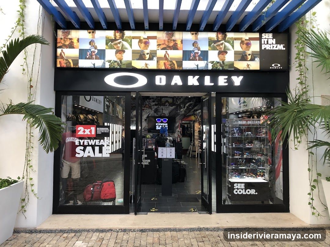 A view of an Oakley retail store entrance. The facade features a black sign with the white Oakley logo and name. Above the sign is a large display of sunglasses models. The glass doors and windows show interior displays, including eyewear on shelves and promotional posters for sales.