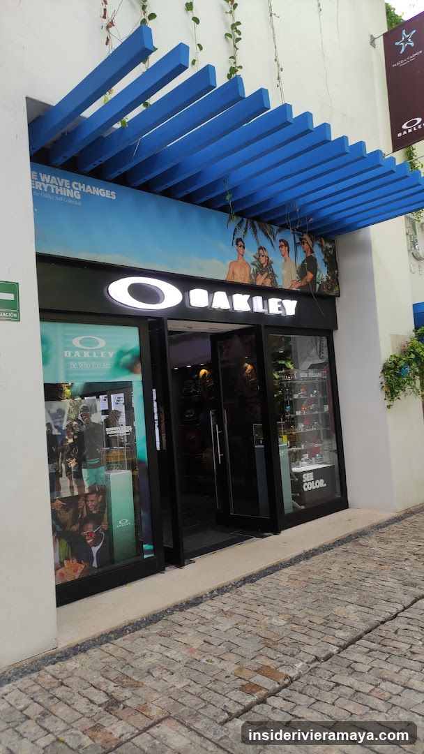 A view of an Oakley retail store entrance featuring a distinctive blue slatted awning. The store has a black facade with a large, illuminated white Oakley logo above the glass doors. Posters are visible in the windows, and the ground is paved with cobblestones.