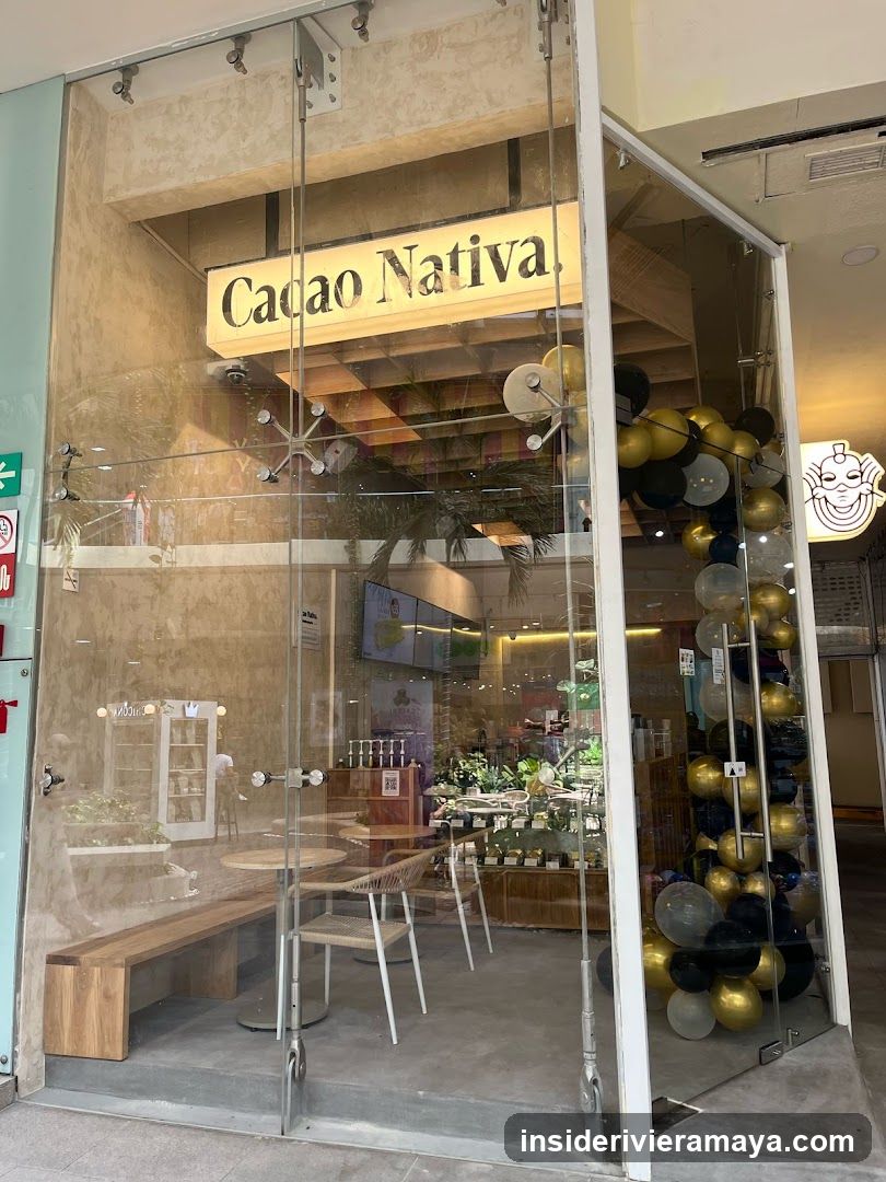 A glass-fronted storefront for a business named 'Cacao Nativa'. The name is displayed on a backlit sign above the entrance. The interior features a wooden bench, tables, and chairs. A decorative column of black, gold, and silver balloons stands to the right of the entrance.