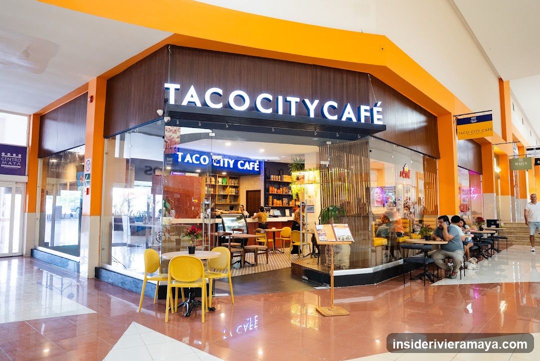 A brightly lit storefront for 'TACO CITY CAFE' located within a shopping mall. The shop features a modern design with orange and dark wood accents, large glass windows, and outdoor seating with yellow chairs. Patrons are seated at tables inside and outside the establishment.