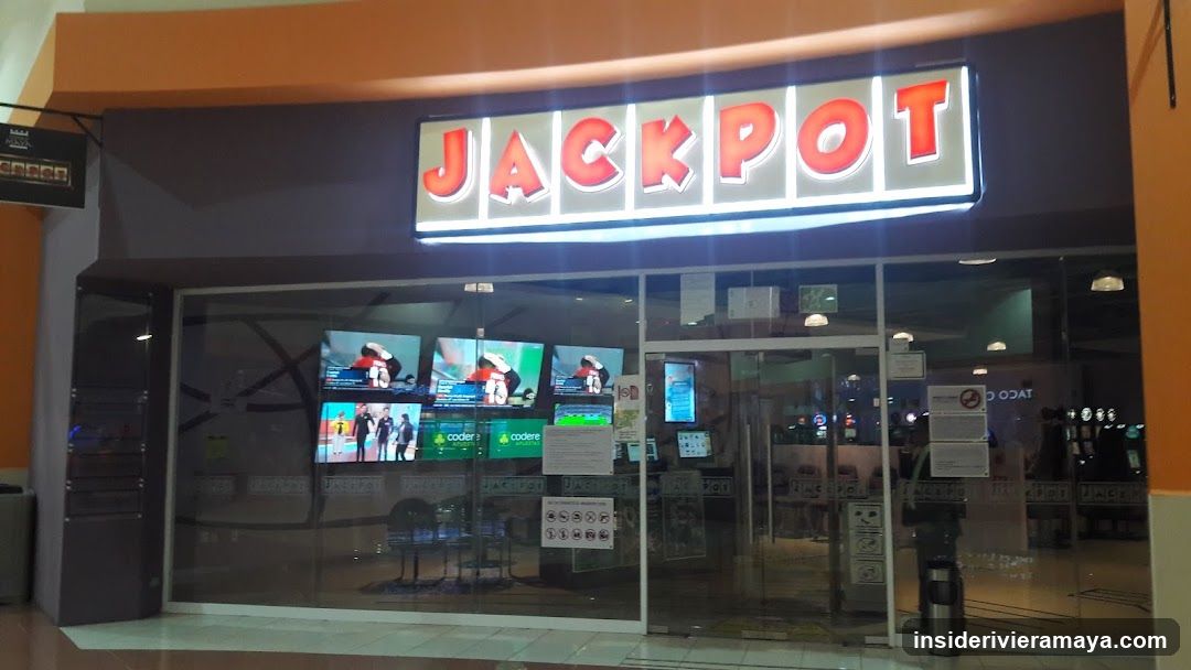 A brightly lit storefront with a large, illuminated sign reading 'JACKPOT' in red letters. The entrance features glass doors and large windows displaying multiple television screens showing sports content. Several informational signs and posters are affixed to the glass.
