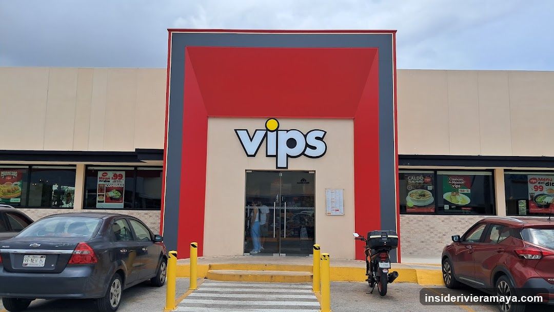 A wide shot of a Vips restaurant storefront featuring a large red and grey facade with the white 'vips' logo centered above the glass entrance doors. Several posters advertising food items are visible in the windows on either side of the entrance. A grey sedan and a red SUV are parked in front, along with a motorcycle.