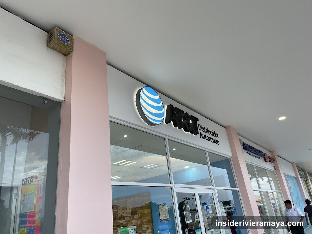 An angled, low-angle shot of a retail storefront with a large, illuminated AT&T logo and the text 'Distribuidor Autorizado' mounted above the entrance. The store features floor-to-ceiling glass windows and doors, reflecting the outdoor environment. A pink pillar supports the white overhang of the building. To the right, other storefronts are visible, and two people are walking on the sidewalk.
