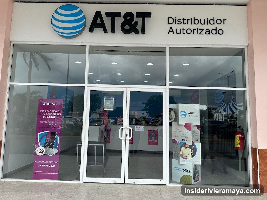 A view of the exterior entrance of an AT&T authorized distributor. The facade features a large white sign with the blue AT&T globe logo and the text 'AT&T Distribuidor Autorizado'. The entrance consists of glass double doors with silver handles, flanked by large glass windows. Several promotional posters are visible inside, including a purple one advertising an AT&T 5G plan for $69 and another for AT&T Business services. A red fire extinguisher is mounted on the wall inside the right window.