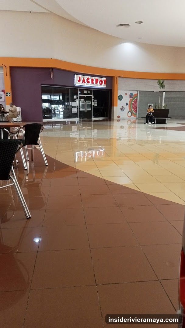 A view of a commercial storefront inside a shopping mall. The store has a purple facade with a prominent red and white sign reading 'JACKPOT' above the glass entrance doors. The floor is tiled in brown and beige, reflecting the overhead lights. To the left, there are black chairs and tables, and to the right, a person is sitting on a bench near a wall with colorful circular graphics.