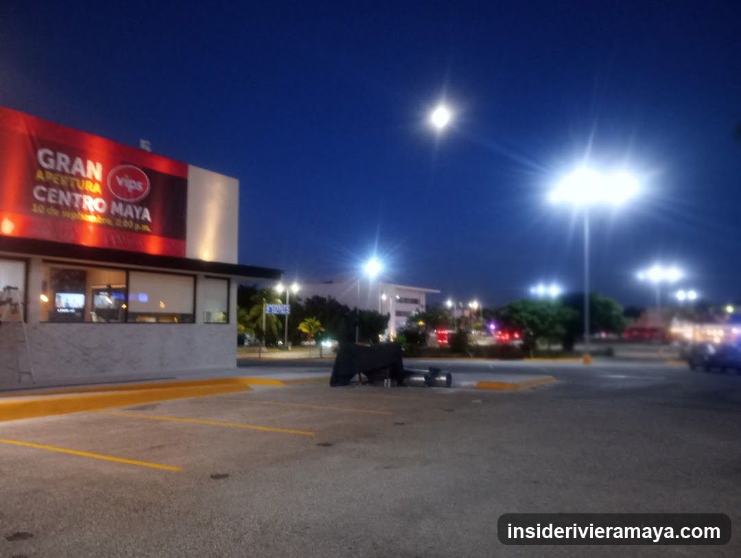 A night view of a Vips restaurant storefront located in Centro Maya. The building features a large red sign with white text announcing a 'GRAN APERTURA' (Grand Opening). The parking lot is paved with asphalt, marked with yellow lines, and illuminated by bright overhead streetlights against a dark blue sky.