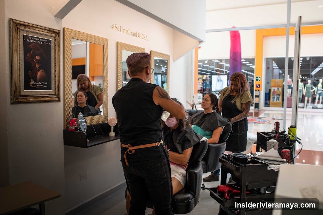 An interior view of a hair salon showing two stylists working on clients seated in black salon chairs. The stylists are wearing black aprons and appear to be cutting or styling hair. The salon has large mirrors with gold frames, a wall-mounted shelf with products, and a framed poster on the left wall. In the background, through glass doors, a shopping mall corridor is visible with mannequins and store displays. A rolling cart with salon tools and supplies is present in the foreground.