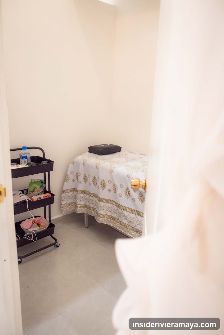 A view through a doorway into a small, clean room containing a massage table covered with a white and gold patterned sheet. A black rolling cart with various supplies, including bottles and bowls, stands next to the table. The room has plain white walls and light-colored tile flooring.