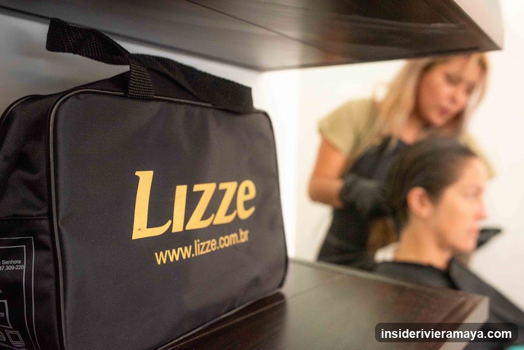 A close-up view of a black fabric bag with the gold text 'Lizze' and the website 'www.lizze.com.br' printed on it. The bag is sitting on a dark wooden shelf. In the blurred background, a hairdresser with blonde hair is styling the hair of a seated female client.