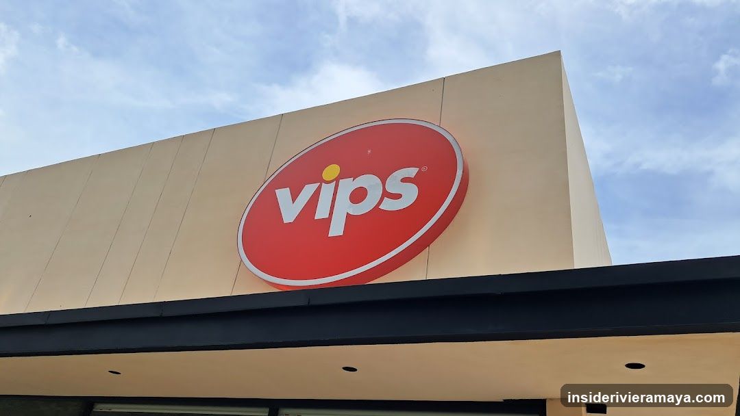 A low-angle shot of a beige commercial building facade featuring a large, red oval sign with the white text 'vips' and a yellow dot over the 'i'. The sign is mounted above a dark overhang against a blue sky.