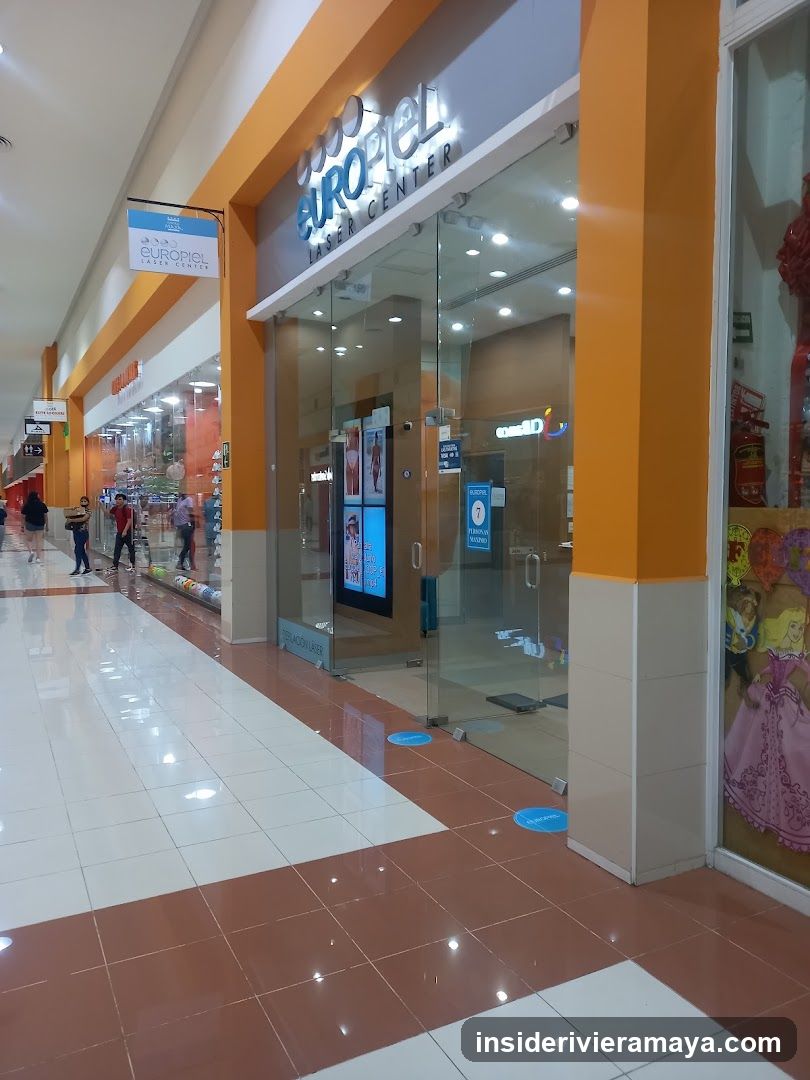 A view of a shopping mall corridor featuring the entrance to a store named 'Euromod Center'. The storefront has large glass doors and windows, framed by bright orange pillars. A sign above the entrance displays the store name in white lettering. Inside, posters are visible on the glass, and blue floor markers are placed near the entrance. Other shoppers can be seen further down the hallway.