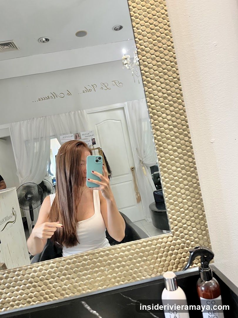 A woman with long brown hair is taking a mirror selfie in what appears to be a hair salon. She is wearing a white tank top and holding a light blue smartphone. The mirror has a textured gold frame. In the reflection, we can see salon equipment like a hair dryer, a white door, and a sign on the wall that reads 'Be La Mode' in reverse. On the counter in the foreground, there are two bottles of hair products, one of which is labeled 'COLLAGEN'.