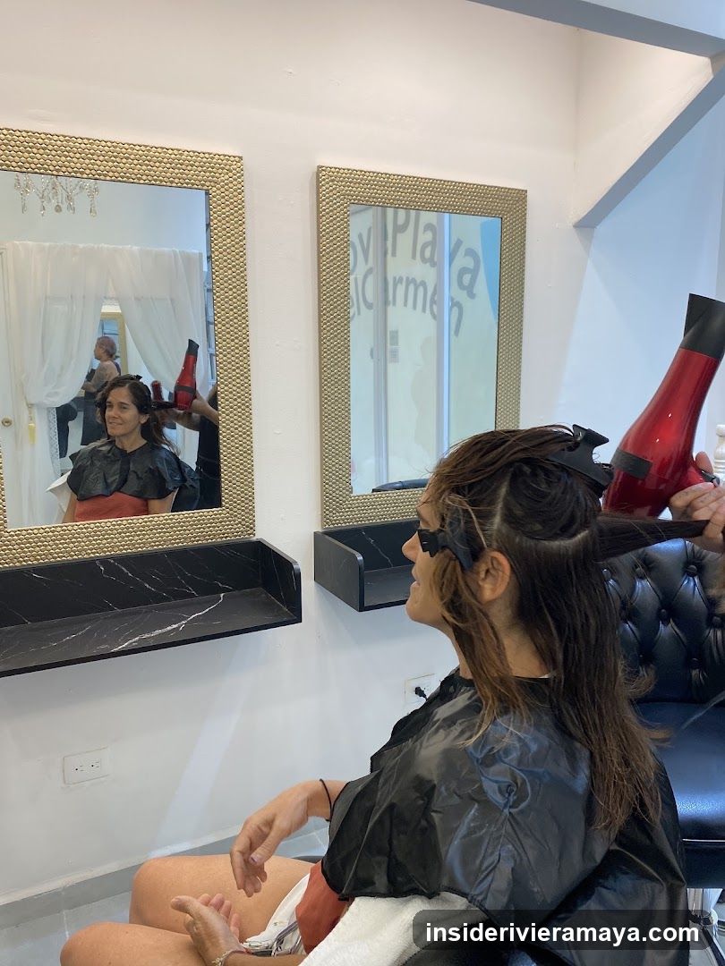 A woman sits in a salon chair wearing a black cape while a stylist uses a red hairdryer on her hair. She is looking into a large mirror with a textured gold frame. The reflection shows her face and the stylist's hands. A second mirror is visible on the wall, reflecting text on a window. The salon has white walls and black marble shelves.