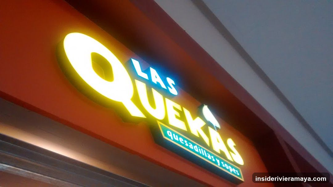 A close-up, angled shot of a brightly lit storefront sign. The sign features the words 'LAS QUEKAS' in large, glowing yellow letters, with 'LAS' in smaller blue letters above. Below the main text, a smaller green sign reads 'quesadillas y sopas'. The sign is mounted on an orange facade.
