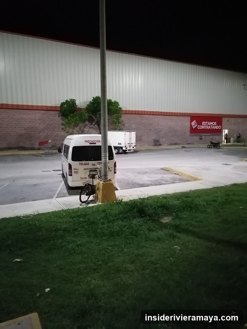 A nighttime view of a large commercial building with a brick lower facade and white siding. A red sign on the wall reads 'ESTAMOS CONTRATANDO' (We are hiring). A white van is parked in the lot, and a bicycle is locked to a light pole in the foreground.