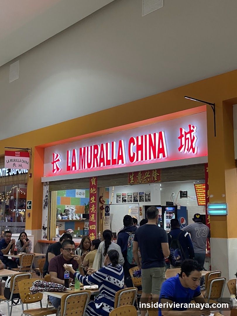 A brightly lit storefront with a large red neon sign reading 'LA MURALLA CHINA' and Chinese characters. The entrance is open, revealing a busy interior with customers seated at tables and staff working behind a counter. Vertical banners with Chinese text hang on either side of the doorway.