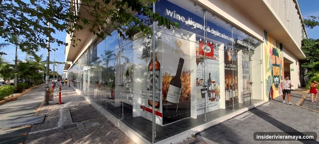 A modern storefront with large glass windows and doors. A blue sign above reads 'wine, liquor & delicatessen'. The windows are covered with large posters advertising various bottles of wine and spirits, including brands like Ventos and Magalão. A colorful mural is visible on the far right wall. People are walking on the sidewalk outside.