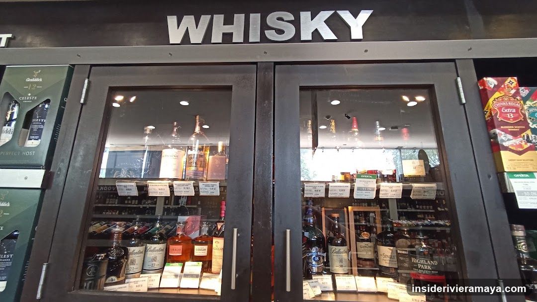 A close-up view of a retail storefront featuring a large, dark-framed glass cabinet labeled 'WHISKY' in bold silver letters. The cabinet contains multiple shelves stocked with various bottles of alcohol, likely whisky, with small white price tags attached to the glass. To the left, a green box for 'Glenfiddich' is visible, and to the right, a box of 'Coca-Cola Extra' sits on a shelf.