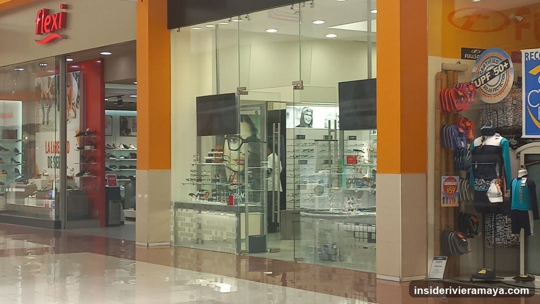 A view of a shopping mall corridor featuring storefronts. On the left, a store with a red 'Flexi' sign displays shoes and accessories. In the center, an optical shop with glass walls and orange pillars showcases eyewear on shelves and mannequins. To the right, a retail display features bags, backpacks, and clothing under a 'UPF 50+' sign.