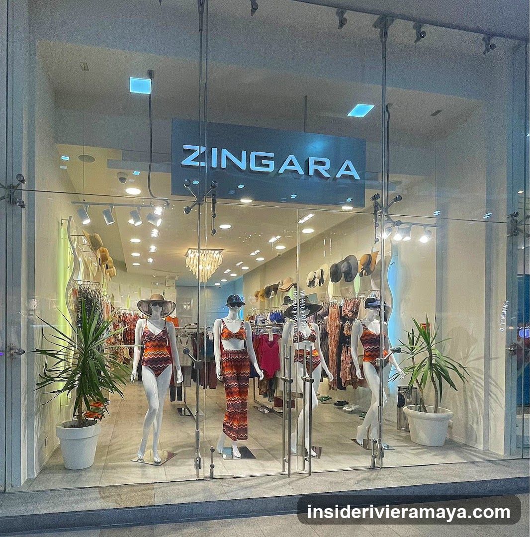 A brightly lit retail store entrance with large glass doors and windows. The store name 'ZINGARA' is displayed in large, illuminated white letters on a dark blue sign above the entrance. Inside, several mannequins are dressed in colorful, patterned swimwear and summer clothing. The interior features hanging lights, racks of clothing, and potted plants near the entrance.