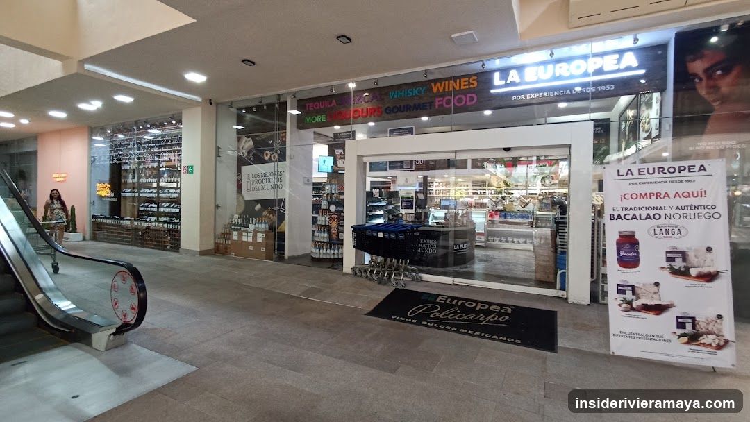 A brightly lit storefront for a business named 'LA EUROPEA' located inside a shopping mall. The entrance features automatic sliding glass doors and a black welcome mat. A large illuminated sign above the store lists categories such as 'TEQUILA', 'WHISKY', 'WINES', 'GOURMETS', and 'FOOD'. To the right of the entrance, a standing poster advertises 'BACALAO NORUEGO' (Norwegian Cod). To the left, a neighboring store selling eyewear is visible next to an escalator.