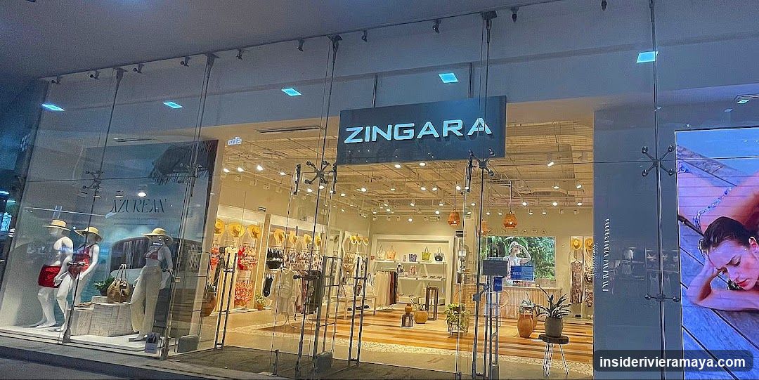 A brightly lit retail store entrance with large glass windows and doors. A prominent black sign with the name 'ZINGARA' in white capital letters hangs above the entrance. Inside, the store features light wood flooring, hanging pendant lights, and displays of clothing and accessories. Mannequins are visible in the window display on the left, and a large digital screen is on the right.