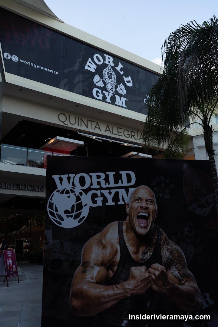 A low-angle shot of a World Gym storefront located in a shopping center called Quinta Alegría. The gym's name is displayed on a large black banner above the entrance, along with social media handles. In the foreground, a large promotional poster features a muscular man shouting, with the World Gym logo.