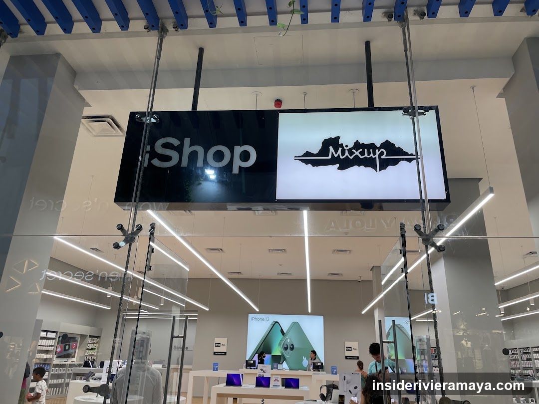 A view of a modern electronics store entrance featuring a large suspended sign reading 'iShop' and 'Mixup'. The storefront is enclosed by glass walls, revealing a brightly lit interior with white display tables, ceiling track lighting, and a large screen advertising an iPhone 11.