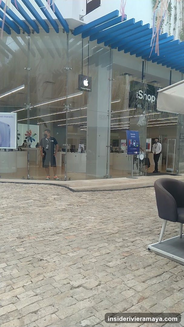 A view of a retail store entrance with a glass facade and blue slatted awning. The store signage reads 'iShop' and features an Apple logo. Inside, staff members are visible standing near the counter.