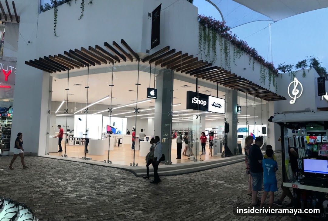 A modern retail store with a white facade and large glass walls. The entrance features a wooden slat pergola roof. A black sign with white text reading 'iShop' and a logo is mounted on the glass. An illuminated musical note symbol is visible on the adjacent wall. Several people are standing outside on a cobblestone plaza, and a small kiosk is visible to the right.