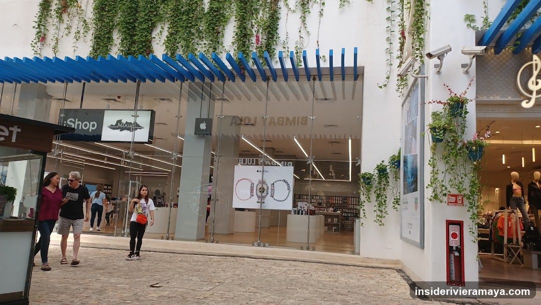 A view of a commercial storefront featuring a prominent blue slatted pergola structure extending over the entrance. The facade is white with hanging greenery. A large glass window displays posters for watches, and signage for 'BIMBA Y LOLA' is visible inside the store. Pedestrians are walking on the paved walkway in front.