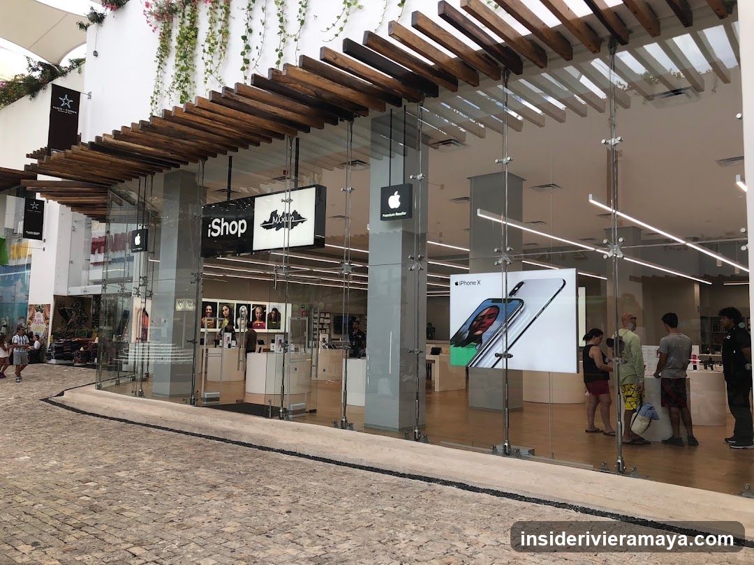 A modern retail storefront with a glass facade and wooden slat pergola. The sign reads 'iShop' and features an Apple logo. Inside, customers are browsing near display tables, and a large poster advertising an iPhone X is visible.