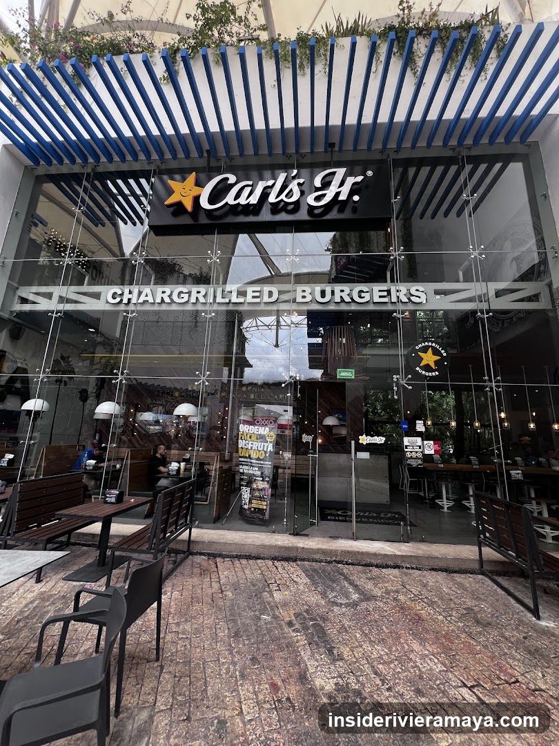 A view of the Carl's Jr. storefront featuring a modern glass facade with blue slat awning. The main signage displays 'Carl's Jr.' with a star logo, and below it, 'CHARGRILLED BURGERS' is printed on the glass. Outdoor seating with dark tables and chairs is visible on a brick-paved patio.