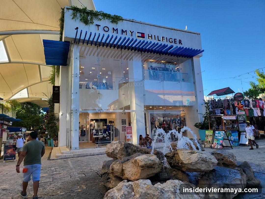 A two-story white building with a glass facade and a blue slatted awning. The sign 'TOMMY HILFIGER' is clearly visible on the upper level. A small rock fountain is in the foreground, and people are walking around the outdoor plaza.