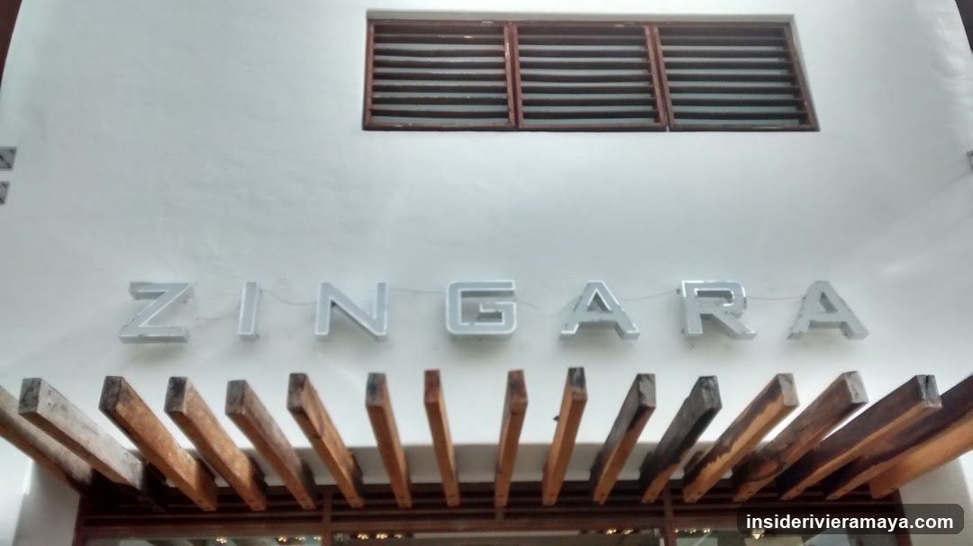 A white building facade featuring the name 'ZINGARA' in large, silver, three-dimensional letters. Below the sign is a wooden pergola structure with horizontal slats. Above the sign is a rectangular window with horizontal louvers.