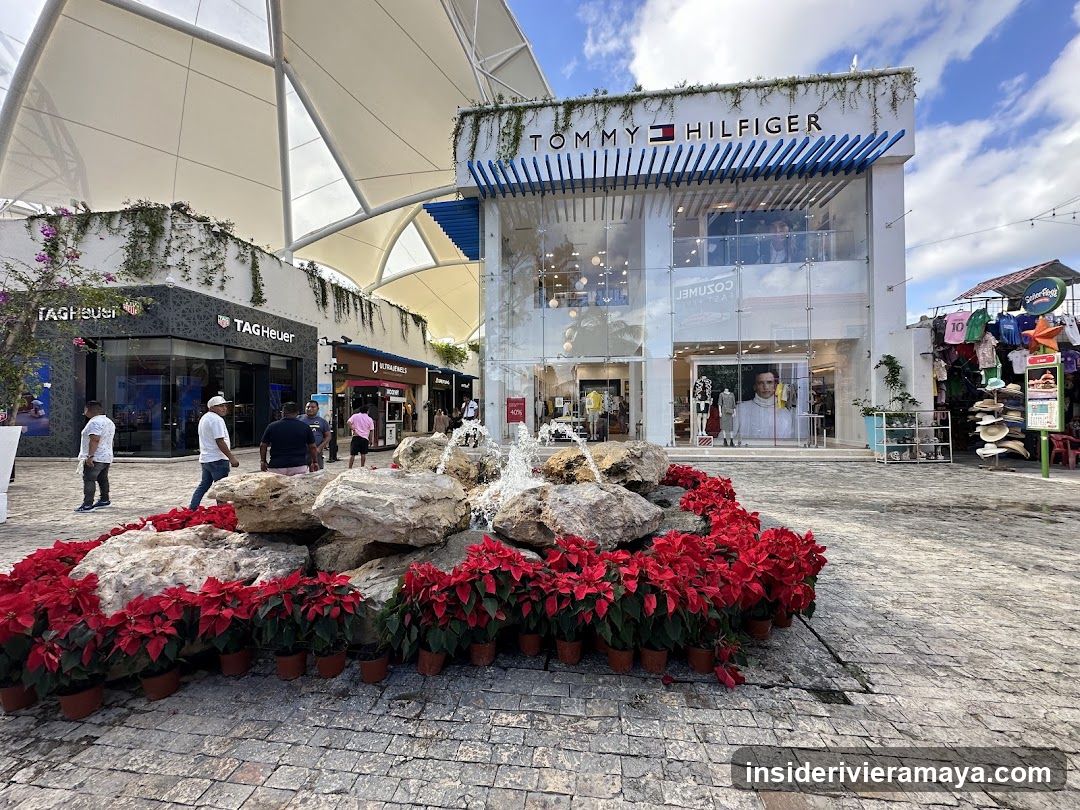 A view of an outdoor shopping plaza featuring storefronts for Tommy Hilfiger and Tag Heuer. A central fountain surrounded by red poinsettias sits in the foreground, with pedestrians walking on the paved ground.