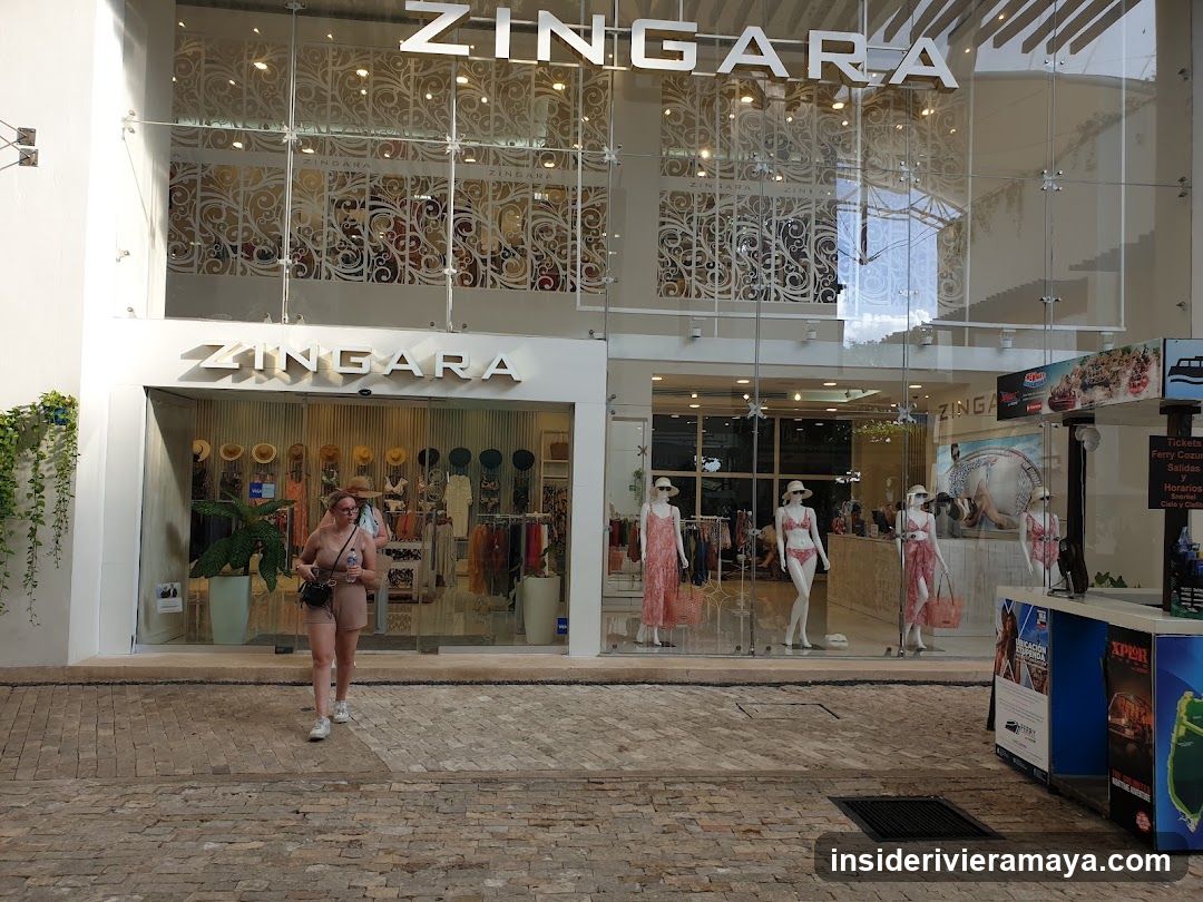 A two-story retail storefront with large glass windows and doors. The store name 'ZINGARA' is displayed in large white letters above the entrance and on the facade. Inside, mannequins wearing swimwear are visible, along with racks of clothing and decorative wall panels. A woman walks past the store on the paved walkway.