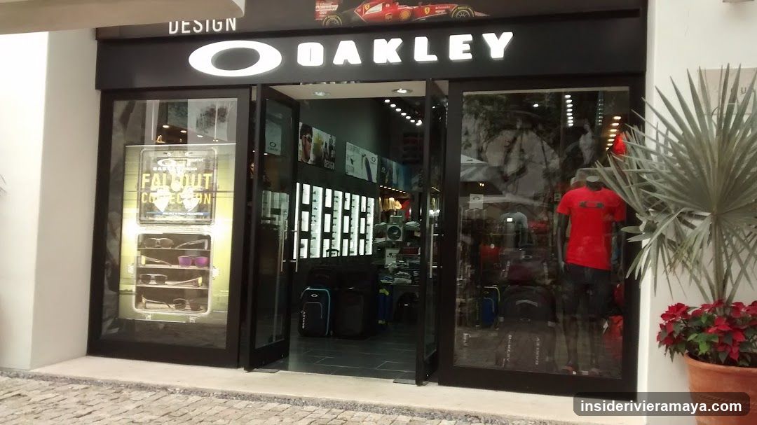 A view of an Oakley retail store entrance. The black facade features large white illuminated lettering for the brand name. Glass doors and windows display merchandise, including sunglasses on wall racks, luggage on the floor, and a mannequin wearing a red shirt. A potted plant with red flowers sits to the right of the entrance.