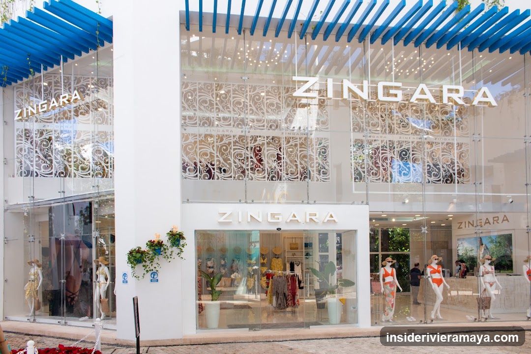 A modern retail storefront with a white facade and large glass windows. The brand name 'ZINGARA' is displayed in large white letters on the upper glass panels and above the central entrance. The store features a distinctive blue slatted pergola structure on the roof. Mannequins wearing swimwear are visible inside the ground floor windows, and decorative white metal screens hang in the upper window area.