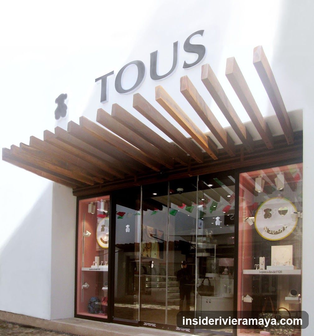 A modern retail storefront with a white facade and a prominent sign reading 'TOUS' in large, dark letters. A distinctive wooden slat canopy extends over the entrance. The store features large glass windows and doors, displaying merchandise inside, including handbags and accessories on shelves and in display cases. A circular logo is visible in the right window.