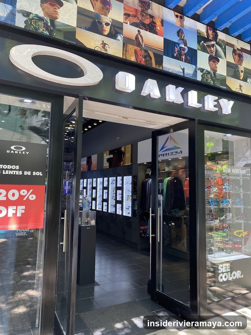 A view of the exterior entrance to an Oakley retail store. The facade features a large, illuminated white 'OAKLEY' sign above the doorway. The entrance consists of glass doors and windows, with a prominent red sign advertising '20% OFF' on the left. Inside, displays of eyewear and clothing are visible.