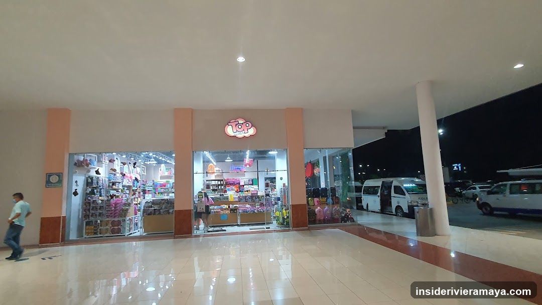 A brightly lit storefront with large glass windows and doors, situated under a covered walkway at night. The facade features peach-colored pillars and a glowing pink logo above the entrance. Inside, shelves stocked with various merchandise are visible. Outside, a white van is parked, and a person is walking past on the left.