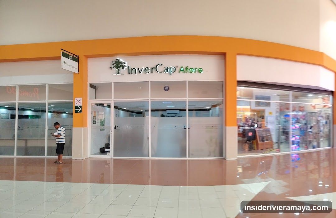 A view of a commercial storefront located within a shopping mall. The store features a prominent orange facade with large glass windows and sliding doors. The business name 'InverCap Afore' is displayed in green lettering above the entrance, accompanied by a tree logo. A man in a striped shirt stands outside the store looking at his phone. To the right, another store with merchandise is partially visible.