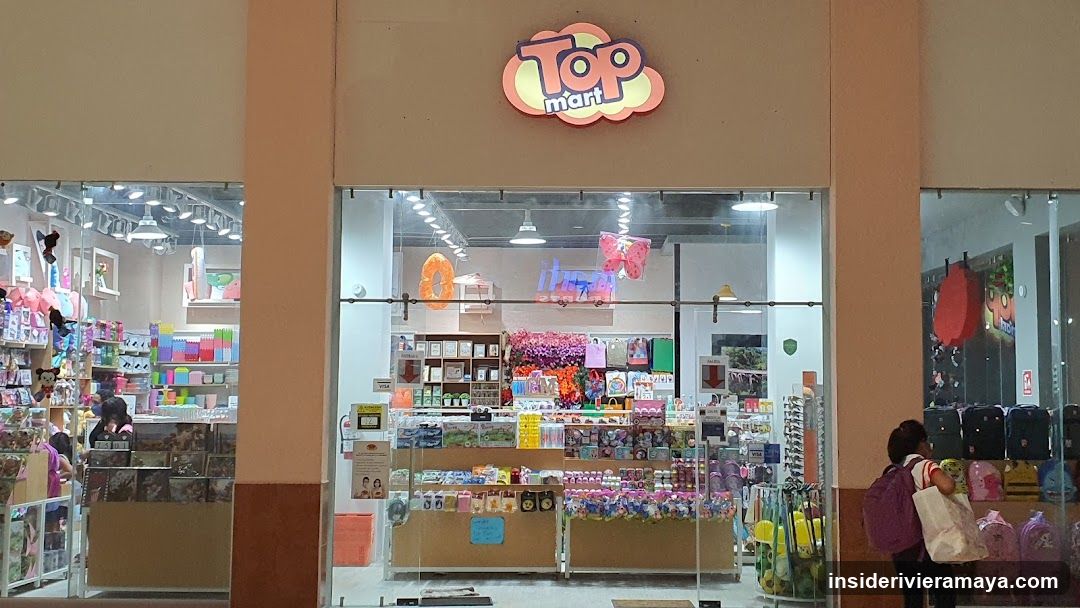 A brightly lit storefront with large glass windows and doors. A colorful, cloud-shaped sign above the entrance reads 'Top mart'. Inside, shelves are stocked with various colorful merchandise, including toys, stationery, and bags. A person with a purple backpack is visible on the right side.