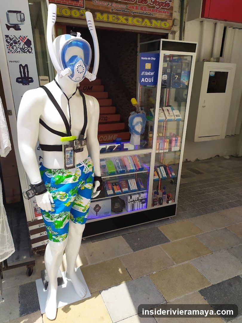 A view of a store entrance featuring a white mannequin wearing blue swim trunks, a black harness, and a blue and white snorkel mask. The mannequin stands on a sidewalk in front of a glass display case filled with small packaged goods. A sign above the entrance reads 'MEXICANO'.