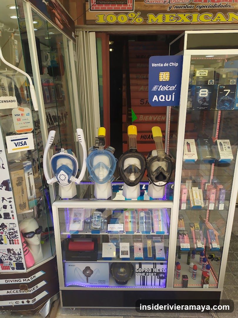 A storefront display featuring snorkeling masks on mannequin heads, a Telcel chip sale sign, and glass cabinets filled with electronics and accessories. A sign above reads '100% MEXICAN'.