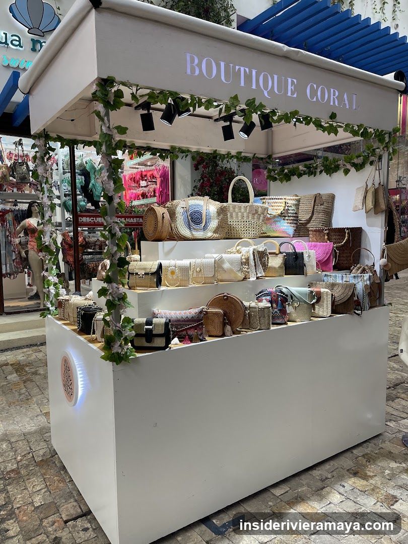 A white outdoor market stall with the sign 'BOUTIQUE CORAL' on the top canopy. The stall is decorated with artificial ivy and displays numerous woven straw bags, handbags, and purses on tiered shelves. A woman is visible browsing in the background store.