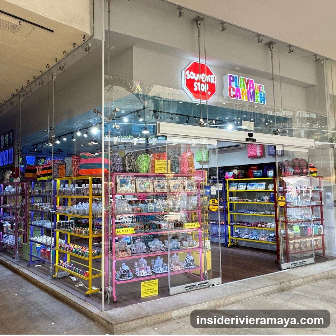 A brightly lit storefront with large glass windows and automatic sliding doors. The facade features prominent signage reading 'SOUVENIR STOP' in a red octagon and 'PLAYA CARMEN' in colorful block letters. Inside, multiple colorful metal shelving units (yellow, pink, purple) are stocked with various merchandise, including bags, bottles, and small souvenirs. Yellow sale signs are visible on the shelves.